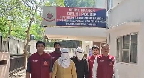 A group of six people, including two with covered faces, stand outside a New Delhi Crime Branch building. Four men wearing red Crime Branch vests surround the two individuals with covered faces, suggesting a formal or official context. The building sign and a tree are visible in the background.