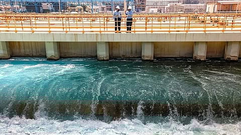 Two workers in hard hats stand on a walkway overlooking a large water treatment facility. Turbulent blue water swirls below, suggesting active filtration.