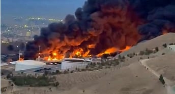 A massive fire engulfs an industrial area, with thick black smoke billowing into the sky. Nearby city lights twinkle under the evening sky, adding contrast.