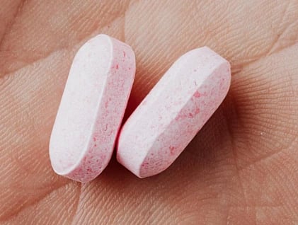 Close-up of two pink oblong pills resting on a palm. The background shows detailed skin texture, suggesting a focus on health or medication.