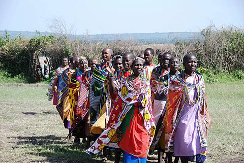 A group of women in colorful traditional Maasai clothing and beadwork stand closely together on a grassy field, smiling and expressing joy under a clear sky.