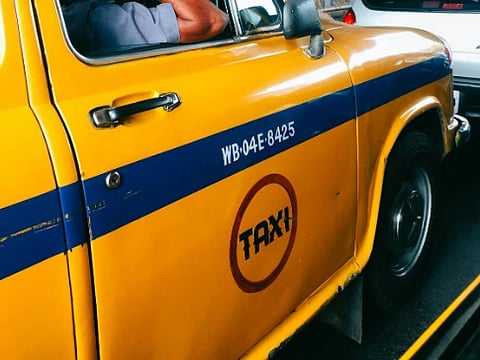 Yellow taxi with a blue stripe, visible license plate WB-04E-8425. "TAXI" in a red circle, driver wearing a blue shirt. Car adjacent in traffic.