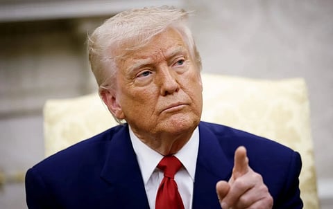 Image of Donald Trump with his classic blue suit and his red tie. His fingers are pointed towards upward. The background is blurred. 