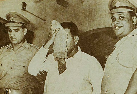 black and white photo of two police officers holding a man in a white kurta who is covering his face with a cloth