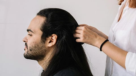 Woman braiding a man's long, dark hair against a light background. She appears focused, while he sits calmly with eyes closed. Casual, serene mood.