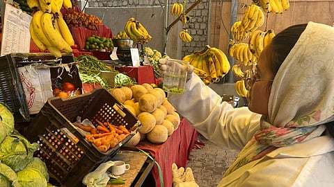 A woman in a headscarf holds up a beaker with some fruits and chemicals in front of a fruit and vegetable stall.