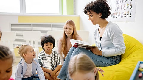 Teacher reading to attentive young children in a classroom, with colorful alphabet poster and bright decor. The mood is warm and engaging.