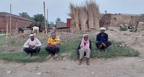 Four men sit crouched on a patch of grass with brick huts in the back