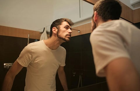 Man in a white t-shirt closely examines his reflection in a bathroom mirror. The setting is calm and introspective, with dark tiles and wood accents.