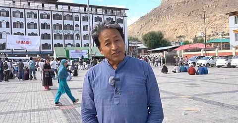 Sonam Wangchuk addressing camera in Ladakh, behind him a building that has a poster of Ladakh, and people walking all around