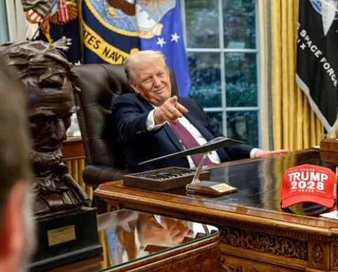 Image of Donald Trump from the White House. he is seated in front of his table. he is pointing towards someone and smiling. there is a cap with text Trump 2028 on his table