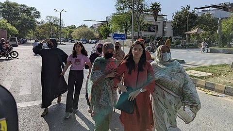 A group of women, wearing traditional and modern attire, confidently cross a sunny urban street. Trees line the road, and buildings are visible in the background.