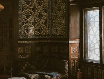 Dimly lit ornate room with patterned walls, arched window, and traditional wooden furniture. The atmosphere is warm and historic.