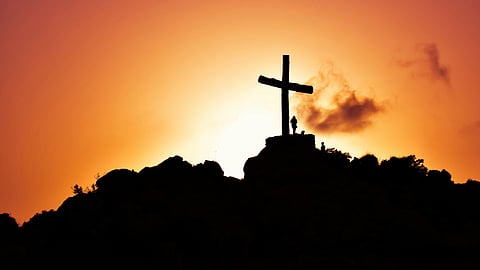 Silhouette of a large cross and a person on a hilltop at sunset, with an orange sky and clouds, conveying a serene and contemplative mood.