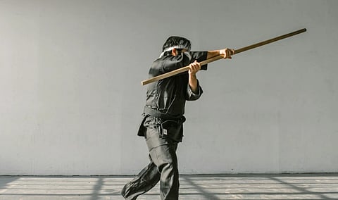A person in black martial arts attire and headband practices with a long wooden staff in a sunlit room, conveying focus and concentration.