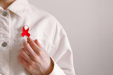 A person wearing a white shirt holds a red awareness ribbon on their chest, symbolizing support or awareness, set against a neutral background.