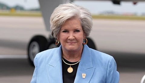 Image of Chief of Staff Susie Wiles in a blue suit and a gold pendant. the background seems to indicate wiles is at a airport. 