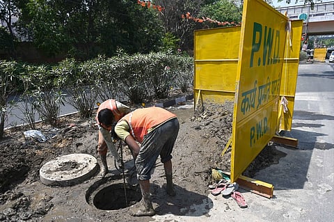 2 sanitation workers bent over an open drain next to a yellow PWD board