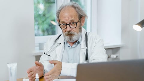 An older Doctor is explaining something in an online meeting, probably to a patient while pointing towards the medicines kept on the table. 