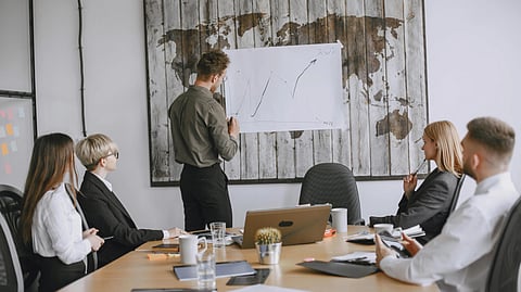 A man presents a line graph to four colleagues in a modern conference room. A world map is on the wall, and laptops and notes are on the table.