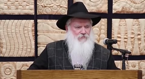 Rabbi Manis Friedman speaks at a podium, wearing a hat, sporting a long white beard