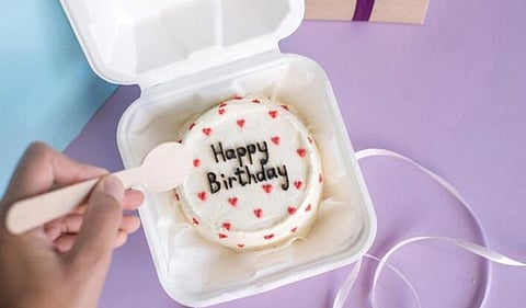 A round cake in a white box is topped with "Happy Birthday" text and red heart decorations. A hand holds a wooden spoon nearby, set on a purple surface.