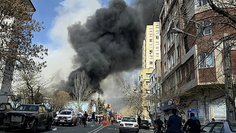 Street scene with thick black smoke rising from a fire, surrounded by onlookers and emergency vehicles, creating a tense and urgent atmosphere.