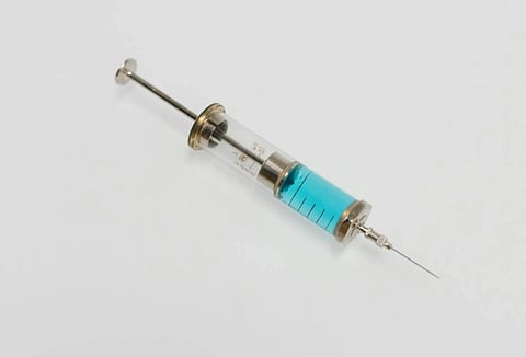 A vintage glass syringe with a metal plunger and needle filled with blue liquid, lying on a white surface. It suggests a medical or scientific context.