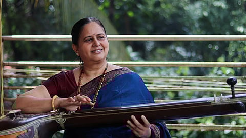 A woman in a blue and burgundy sari plays a veena outdoors. She smiles softly, surrounded by greenery, conveying a serene and joyful mood.