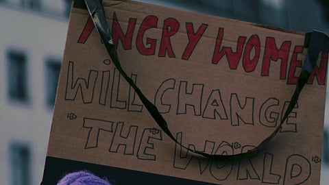 Person in a purple hat and scarf holds a protest sign reading, "Angry Women Will Change the World." The scene conveys empowerment and determination.
