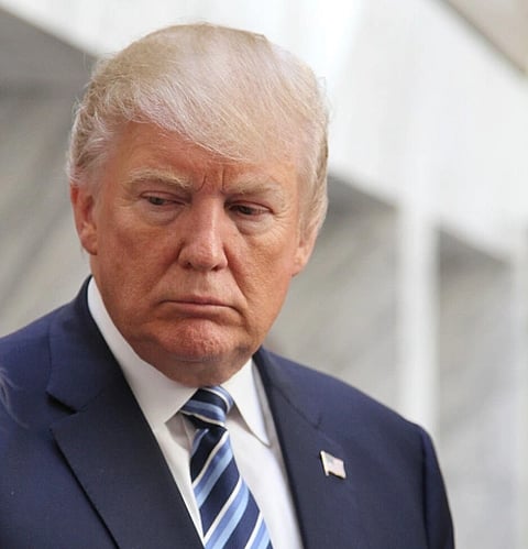 Image of Donald trump in his dark blue suit and badge of american flag. he is looking downwards and has frown on his face. He is at the DC Trump Hotel