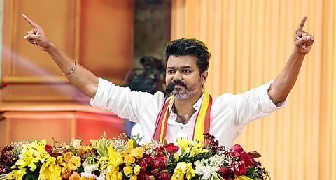 TVK chief Vijay stands with arms raised, smiling confidently, behind a podium adorned with vibrant flowers