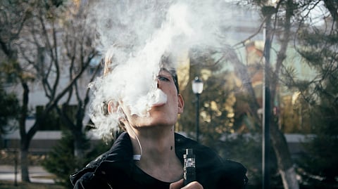 Person vaping outside, exhaling thick white vapor obscuring face, surrounded by leafless trees and blurred urban background, evoking a relaxed mood.