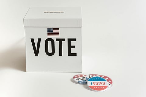 A white ballot box with "VOTE" and a small American flag printed on it. Nearby lie "I Voted" stickers in red, white, and blue, symbolizing civic participation.