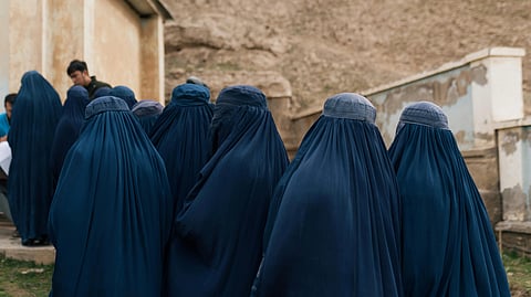 A group of women wearing deep blue burqas stand together outdoors, facing away. The background features an aged wall and a grassy area, creating a somber tone.