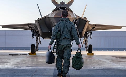 A pilot in a flight suit walks toward a parked fighter jet, carrying bags in each hand. The scene conveys a sense of duty and readiness.
