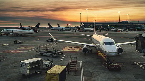 Airplanes are parked on an airport tarmac at sunset, creating a peaceful and busy scene. The sky is a blend of orange and blue hues.