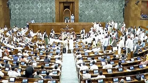 A large parliamentary chamber filled with people in formal attire. Many are seated at desks with laptops, while others stand and converse, creating a bustling atmosphere.