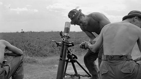 Three shirtless soldiers operate a mortar in a field under a cloudy sky. The scene conveys focus and teamwork in a military setting.