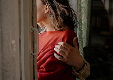 A person in a red sweater looks out a window, with another person's arm gently embracing them, conveying warmth and intimacy. Soft lighting enhances the cozy atmosphere.