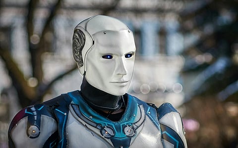 A humanoid robot with a white face and blue-lit eyes stands against a blurred outdoor background. Its expression is neutral and futuristic.