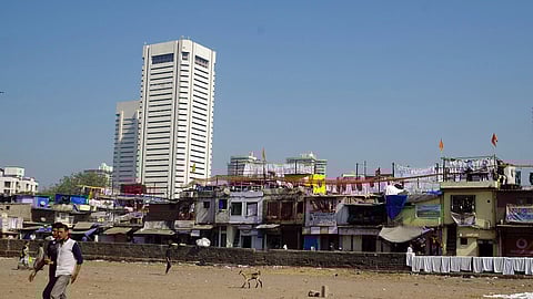 a white building on the left towers over slums on the right, with sand in the foreground