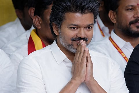 Tamil Nadu Election 2026|TVK leader Vijay in a white shirt with folded hands in a gesture of respect, smiling softly. He is seated among others,