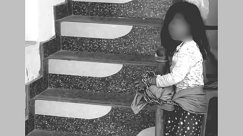 A child wearing a long-sleeve shirt and red pants with white patterns is tied to a staircase railing with rope. The setting appears somber and concerning.