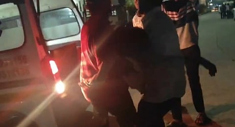 Three people assisting each other under dim street lighting near an open vehicle, suggesting urgency. The mood is tense and serious.