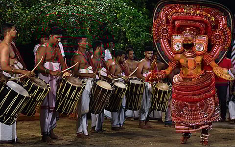 Image of Theyyam of Kerala by Shagil Kannur 2024. A traditional Theyyam dancer in elaborate red costume and face makeup performs energetically before a group of drummers, creating a lively scene.