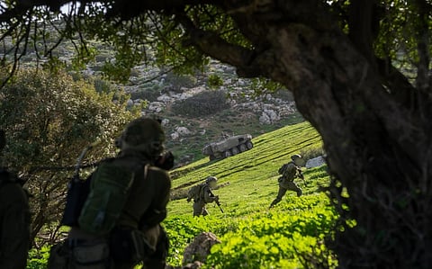 Image of IDF 162nd Division on the Israeli-Lebanese border, Operation Roaring Lion 2026. Soldiers in military gear run through a grassy, hilly landscape, partially shaded by trees, with a military vehicle on a slope in the background.