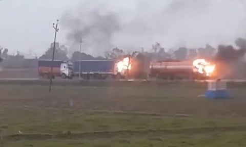 Trucks on a roadside, engulfed in flames and smoke against a gray sky. The scene conveys urgency and chaos. Green fields surround the area.