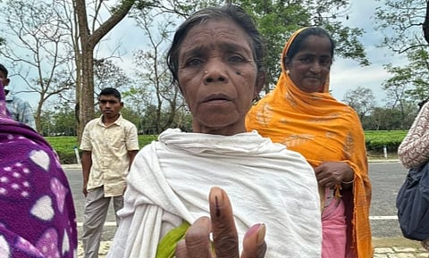 An elderly woman stands foreground, showing an inked finger, symbolizing voting, with a serious expression. Two others stand behind, outdoors on a tree-lined road.