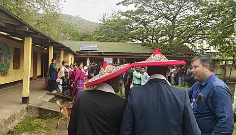 A group of people, including two men in suits with red traditional hats, gathers outside a building. A goat grazes nearby, and a tree and mountains are in the background. The scene feels lively and culturally rich.